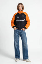 Woman wearing orange and black McLaren racing jacket with sponsor logos, paired with jeans from revnhaus