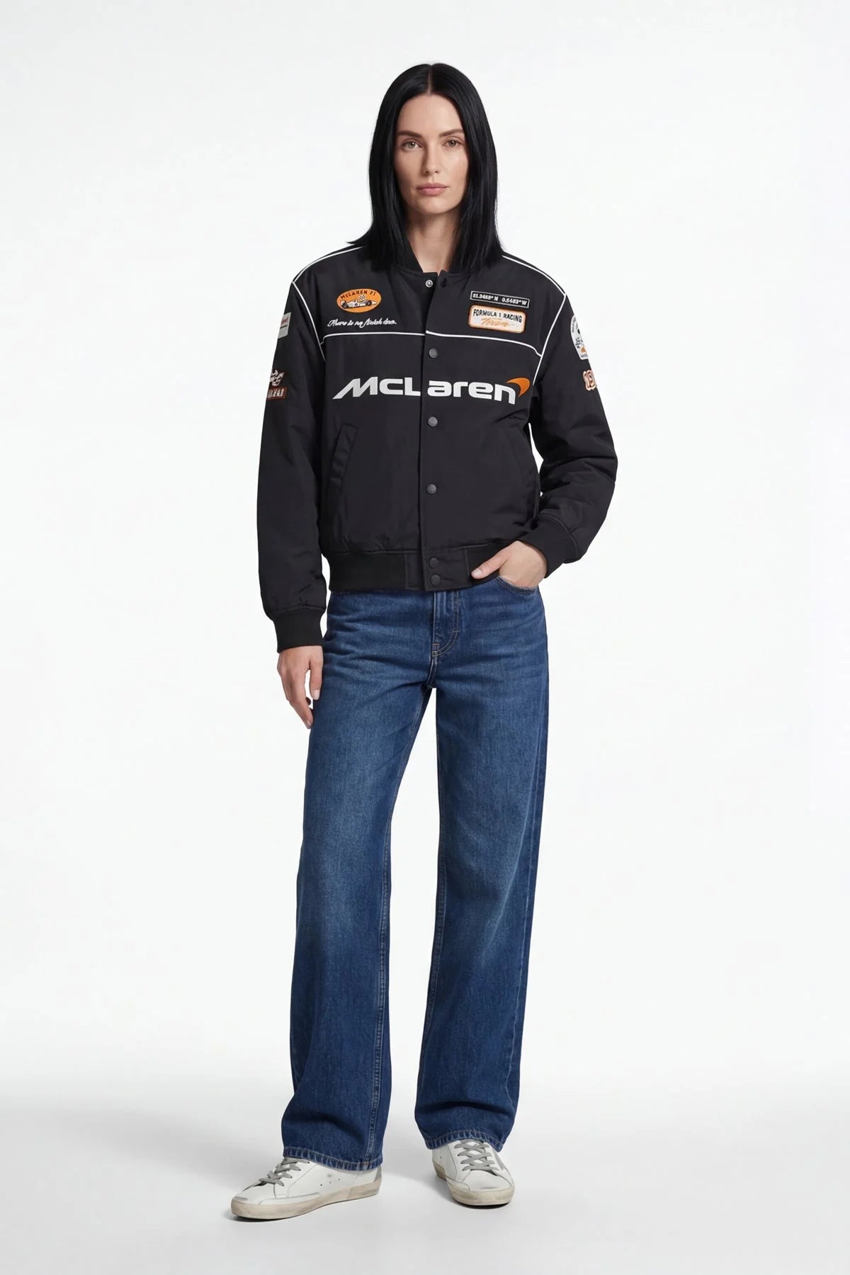 Woman wearing black McLaren racing bomber jacket with patches, blue jeans, white sneakers from revnhaus