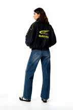 Black Subaru World Rally Team jacket with yellow logo, worn by woman in jeans, from revnhaus